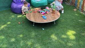 A circular wooden table with toys and a green bean bag chair in a garden area with artificial grass and a green wall. - Bellavista Stone House (Urgup)