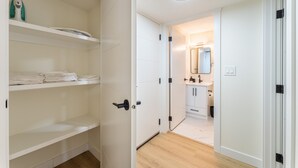 Un couloir mène à une salle de bain équipée d’un miroir et d’un lavabo. Sur le côté gauche, on trouve des étagères blanches avec des serviettes pliées et un fer à repasser.