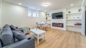 Un salon moderne avec un canapé d’angle gris, une table basse blanche, un coin repas avec une table et des chaises blanches, un téléviseur à écran plat fixé au mur et une cheminée.
