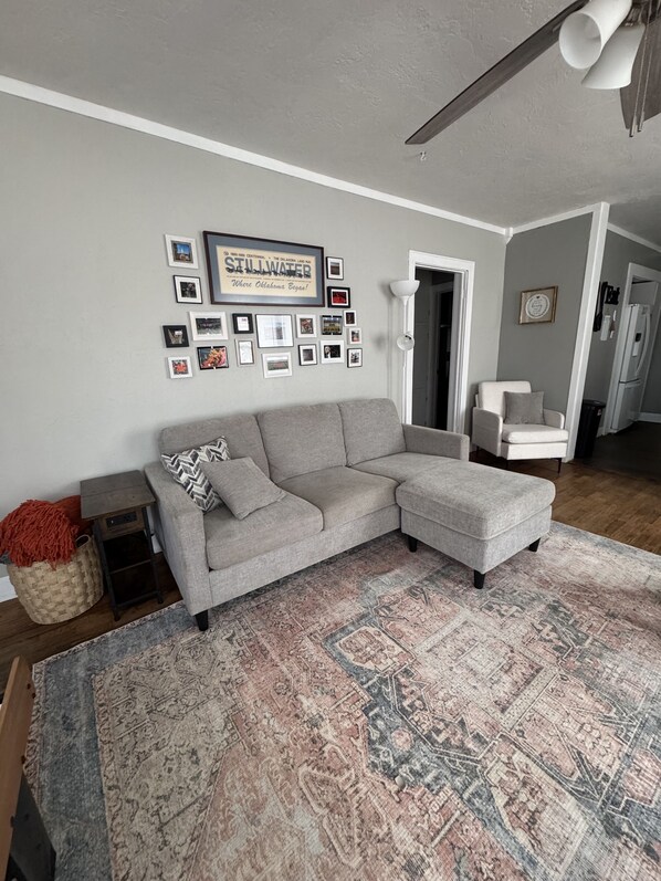 A living room with a grey sectional sofa, a patterned rug, and a wall decorated with framed photos and a 'Stillwater' sign. - Comfortable and Spacious Home close to OSU Campus (Stillwater)