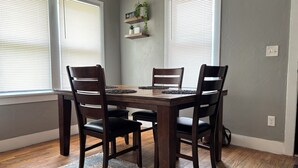 A dining area with a wooden table and four chairs, two windows with blinds, a rug, and a shelf with plants and a sign. - Comfortable and Spacious Home close to OSU Campus (Stillwater)