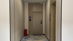 A hallway with a wooden door at the end, a red trash bin to the left, and a light fixture on the ceiling.