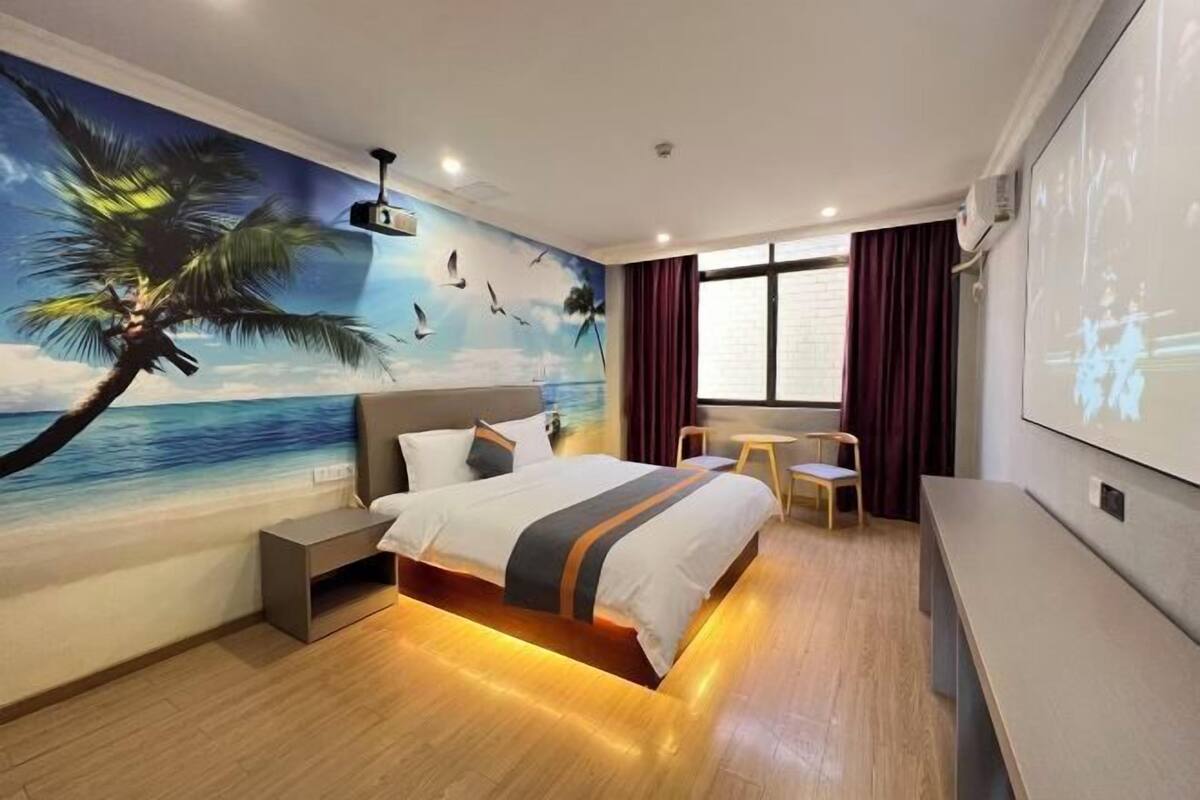 A hotel room with a bed, a desk, a chair, a TV, and a mural of a beach scene.