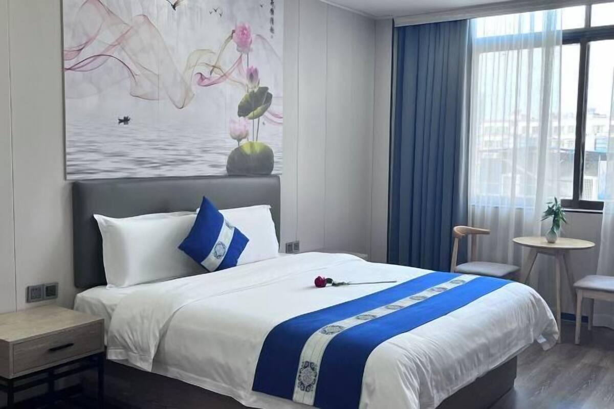 A hotel room with a bed, a small table, a chair, and a window with curtains.