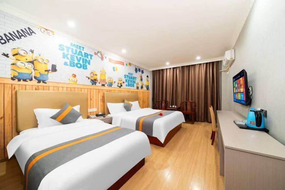 A hotel room with two beds, a desk, a TV, and a wall mural featuring Minions.