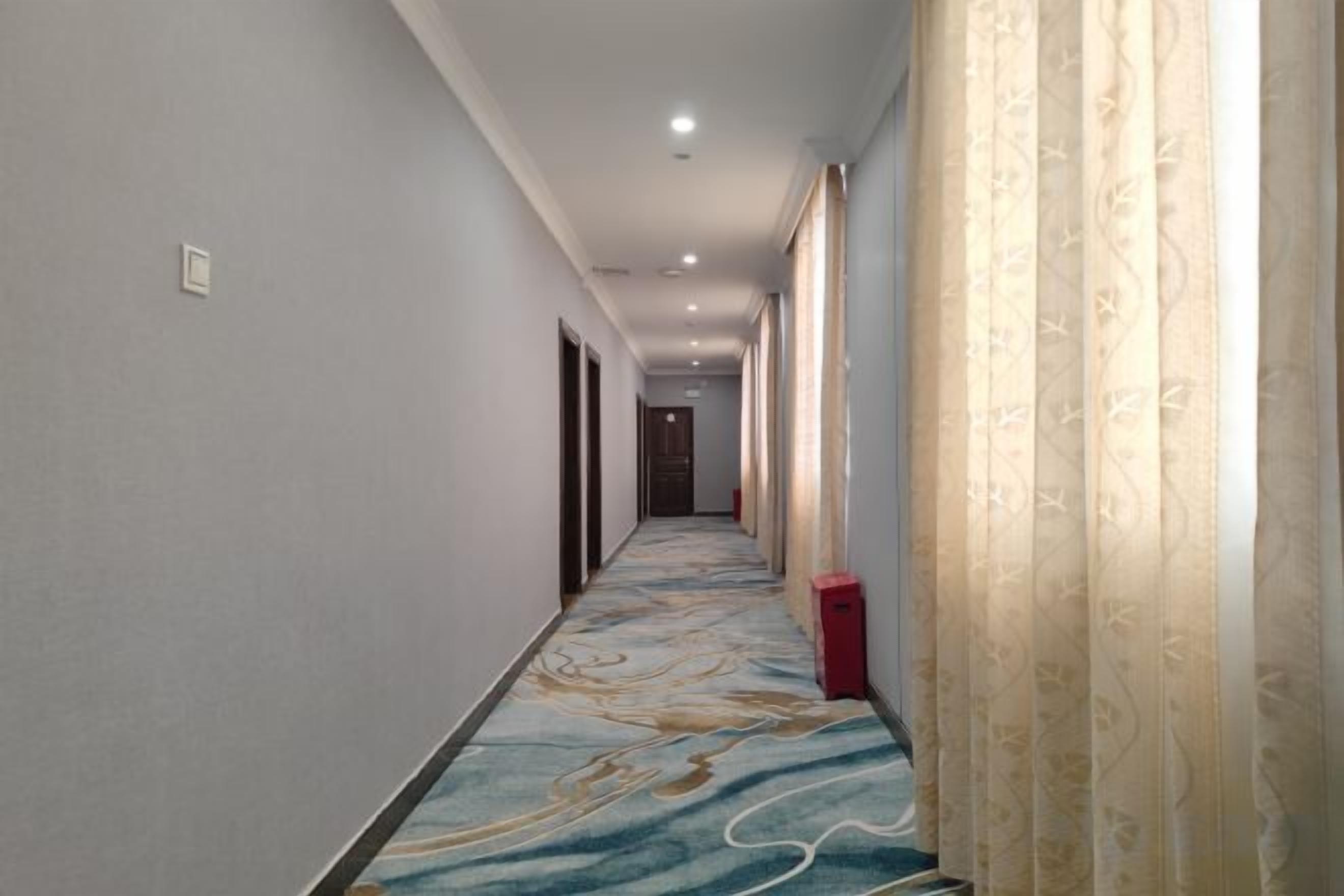 Pasillo de hotel con alfombra estampada, puertas a ambos lados y un bote de basura rojo.