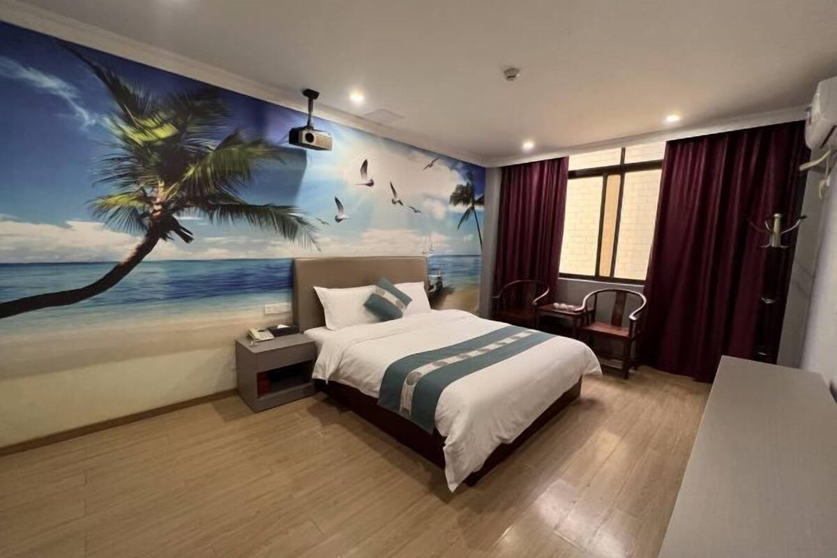 A hotel room with a bed, a nightstand, a desk, a chair, a window with curtains, and a wall mural of a beach scene.