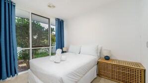 Ein Schlafzimmer mit einem großen Bett, einem fenster mit Vorhängen, einem Nachttisch mit Lampe und einer Holzaufbewahrungseinheit.