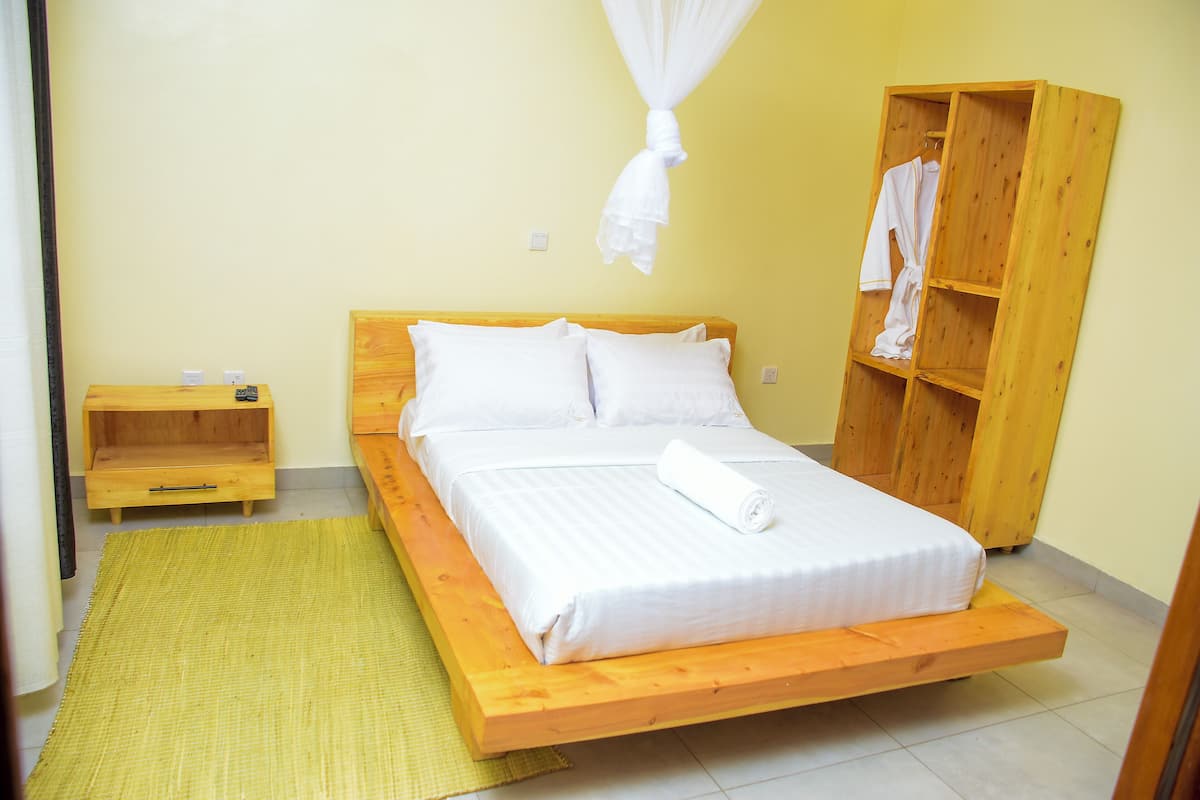 Ein Einzelbett mit Holzrahmen, weißer Bettwäsche und einem weißen Handtuch darauf. Ein Holzschrank mit einem daran hängenden weißen Handtuch. Ein kleiner Nachttisch mit einer Lampe steht neben dem Bett.