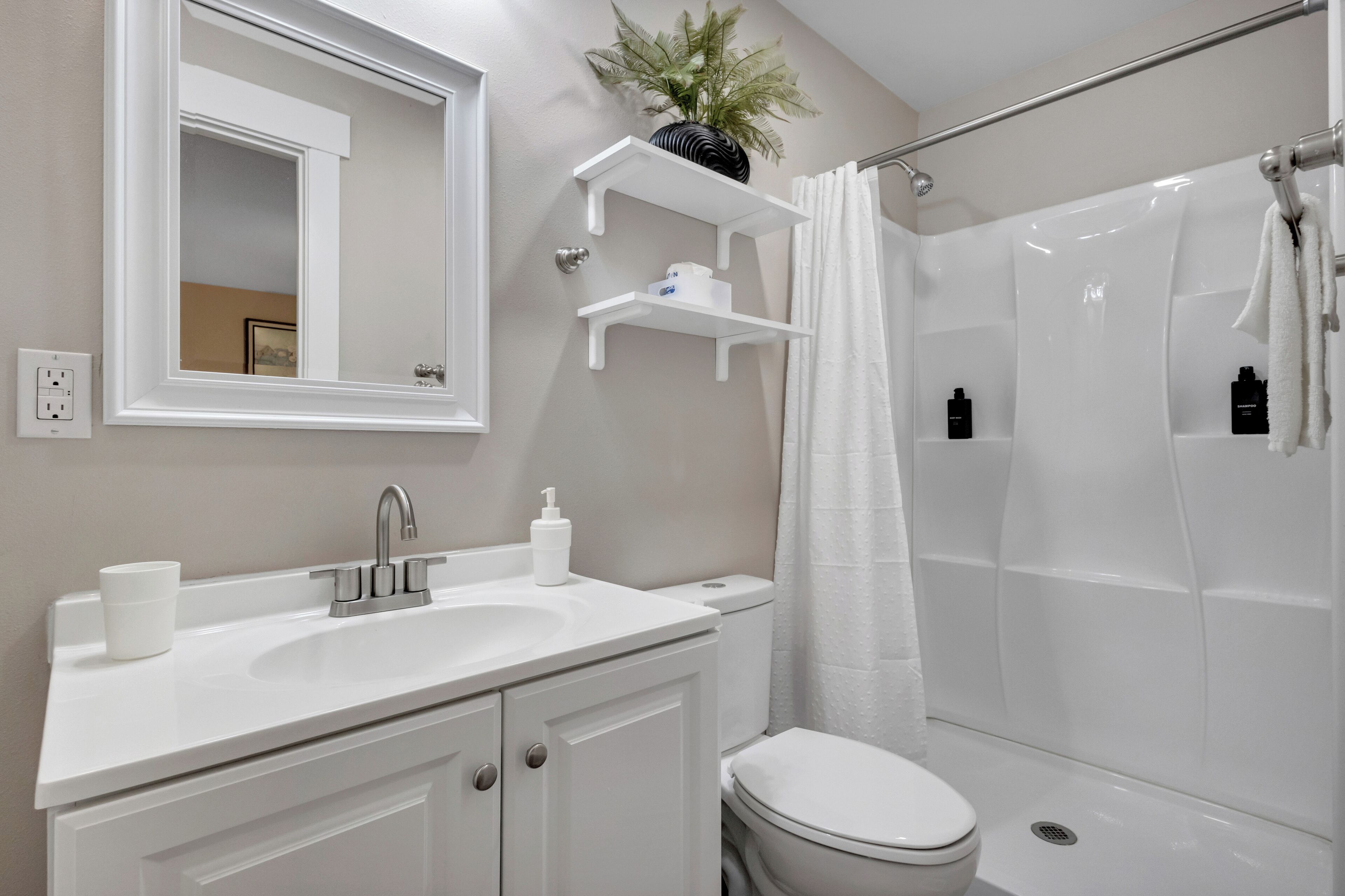 Une salle de bain moderne avec un lavabo blanc, un miroir et des toilettes. On y trouve des étagères blanches au-dessus du lavabo et un rideau de douche.