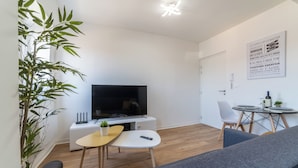 A modern living room with a flat-screen TV, a small table with a plant, a grey sofa, and a dining area with a table and chairs. - The Sweet yachtsman (Mulhouse)