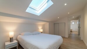 Een slaapkamer met dakraam, een bed met witte beddengoed, twee nachtlampjes en een houten vloer.