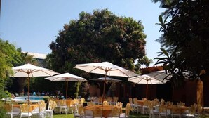 Een evenementenruimte buiten met ronde tafels en stoelen, omringd door parasols, en een zwembad op de achtergrond.