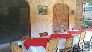 Een buitenontbijtgebied met tafels bedekt met witte tafelkleden en rode tafelrunners, omringd door rieten stoelen.