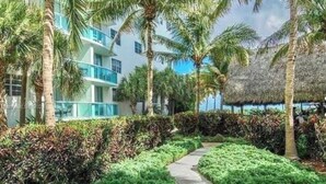 A pathway flanked by lush greenery and palm trees, leading to a building with balconies and a thatched roof structure. - Chic and modern 1-bedroom apartment with 0cean view, WiFi in vibrant Hollywood (Hollywood)