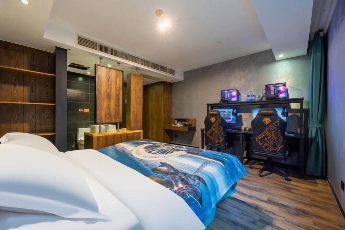 Egy modern szállodai szoba, amelyben egy nagy ágy, egy két monitorral felszerelt íróasztal és egy gamer szék található.