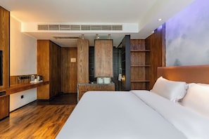 A modern hotel room with a large bed, wooden paneling, a built-in desk, and a seating area with a view of the city.