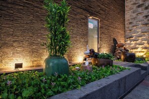 Een goed verlicht buitengebied met een stenen muur, een grote groene plantenbak met planten en een zithoek met een tafel en stoelen.