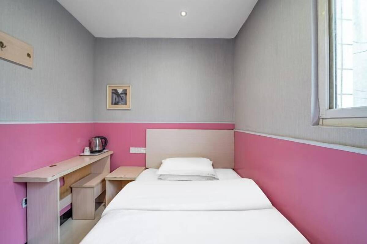 A single-bed room with a pink and grey color scheme, a bedside table with a kettle, and a framed picture on the wall.