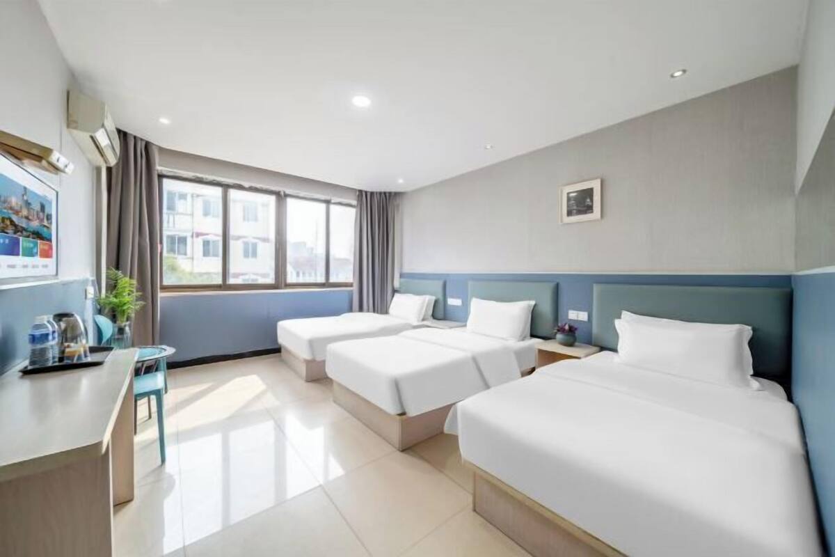A hotel room with two beds, a desk, a chair, a window with curtains, and a small plant.