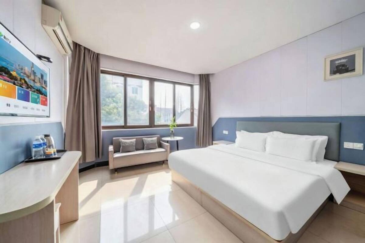 A hotel room with a bed, a sofa, a small table, a window, and a wall-mounted TV.