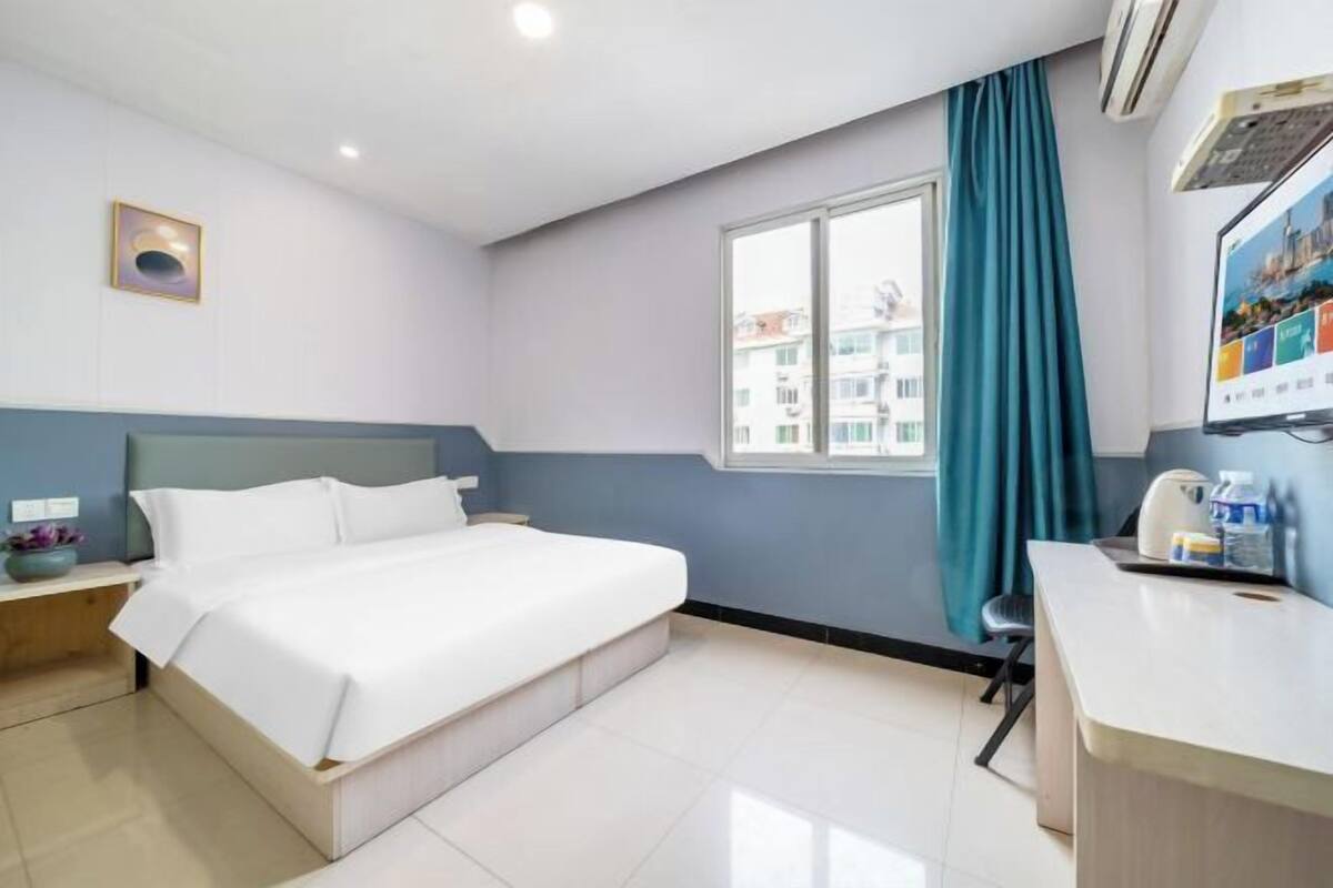 A hotel room with a bed, a desk, a window with curtains, a wall-mounted air conditioner, and a framed picture.