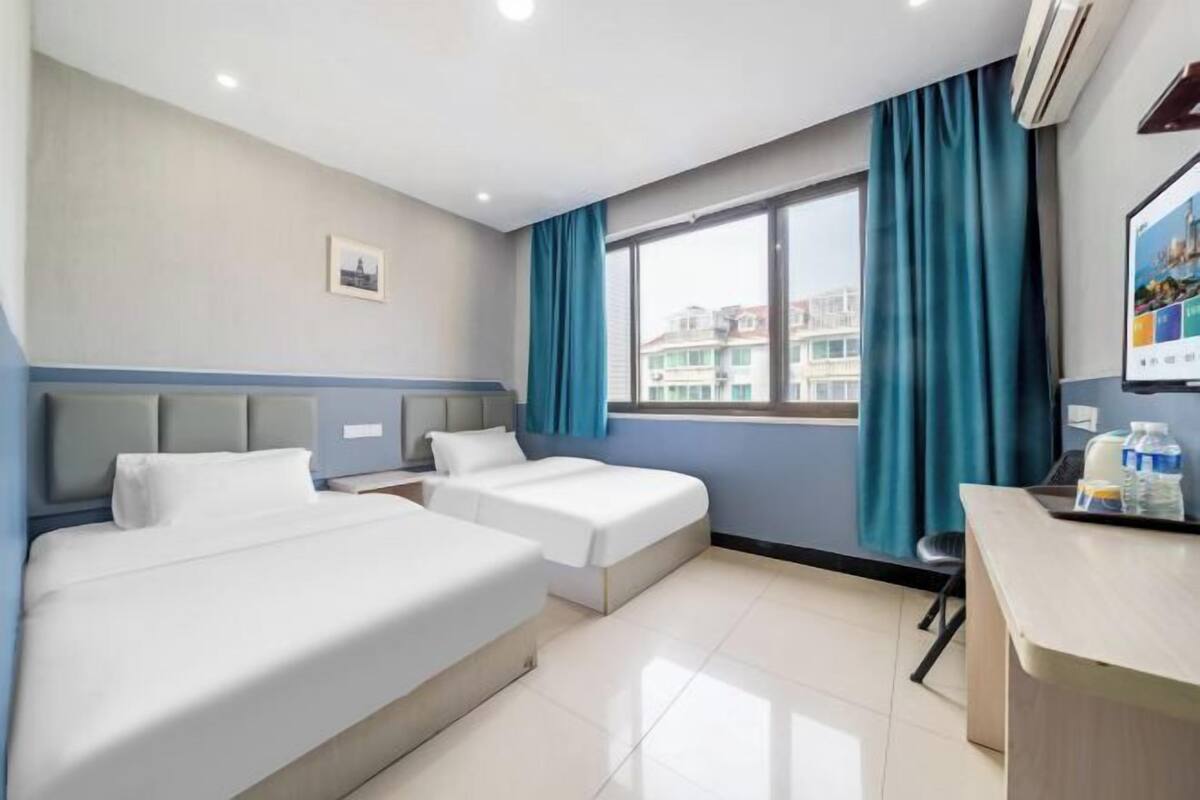 A hotel room with two beds, a desk, a chair, a window with curtains, and an air conditioning unit.