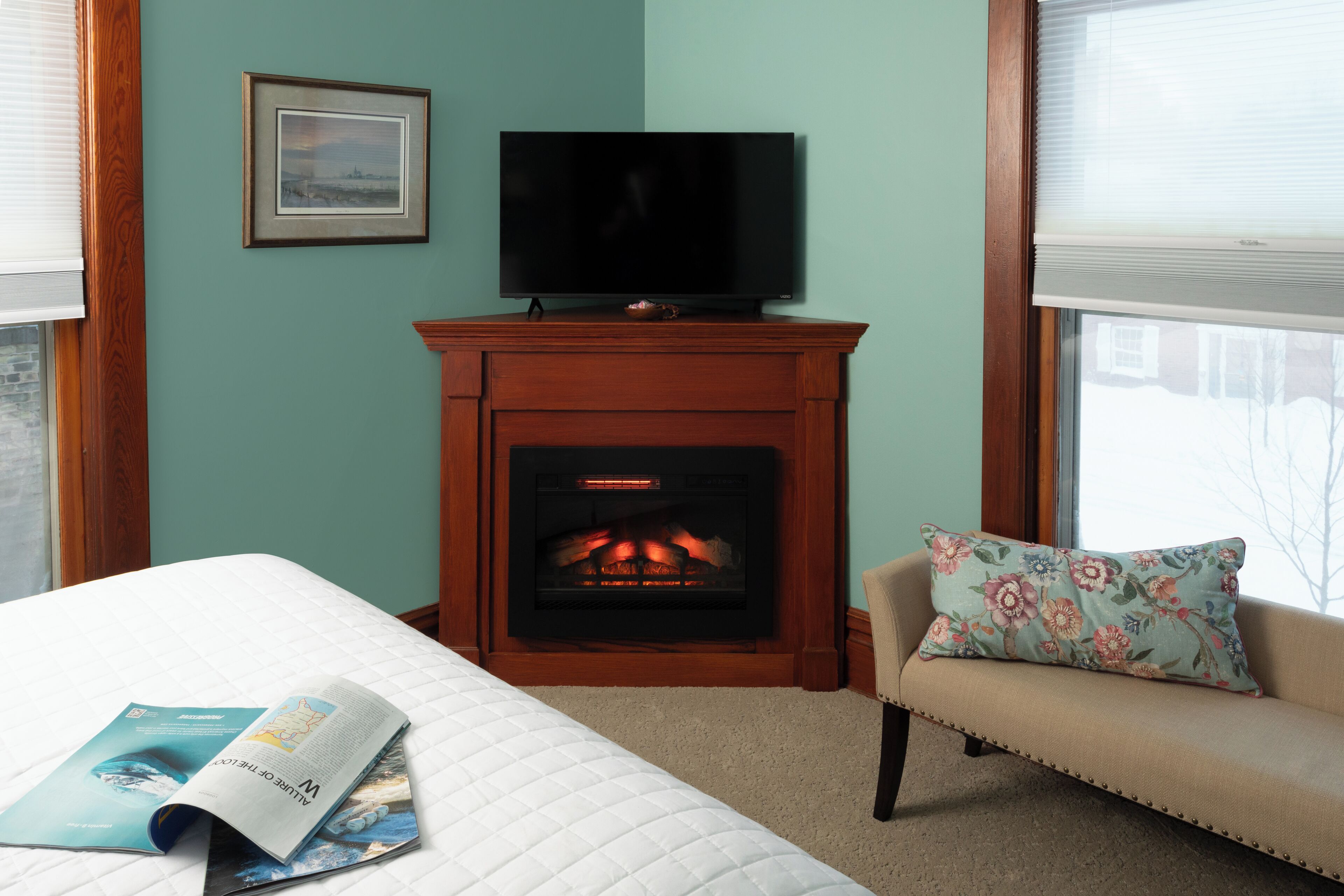 Una habitación con chimenea, un televisor en la pared, un sofá con un cojín floral y una cama con una revista encima.