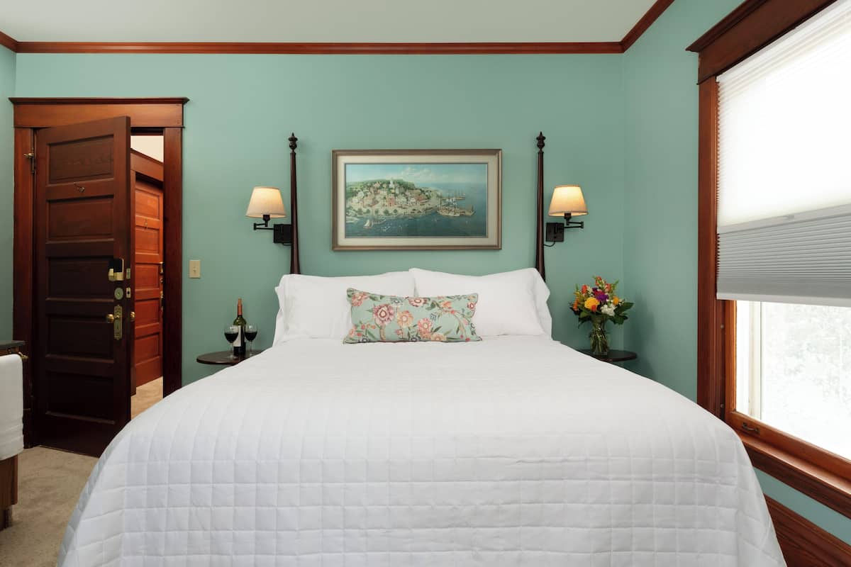A bedroom with a bed, two wall-mounted lamps, a painting, a window with blinds, and a door.