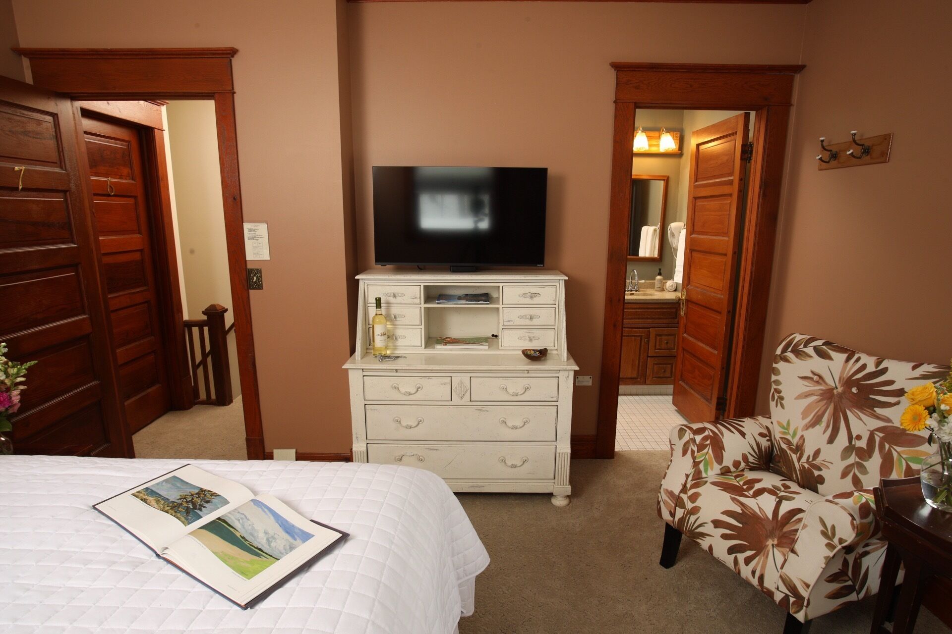 Een hotelkamer met een bed, een witte dressoir met een televisie, een bloemen fauteuil en houten deuren.