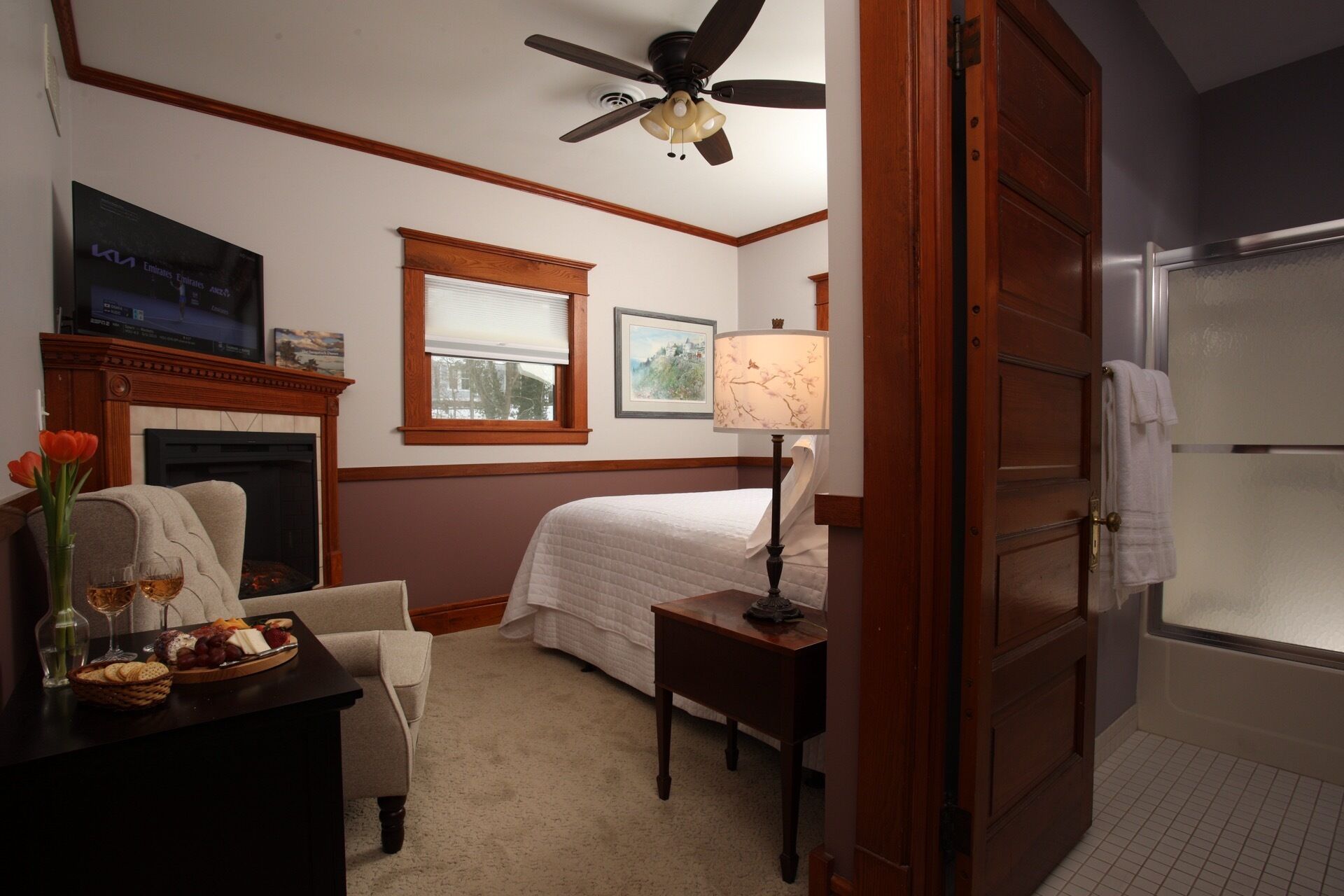 Una habitación de hotel con una cama, una silla, una mesa con aperitivos, una chimenea, un ventilador de techo y un baño con perchero.