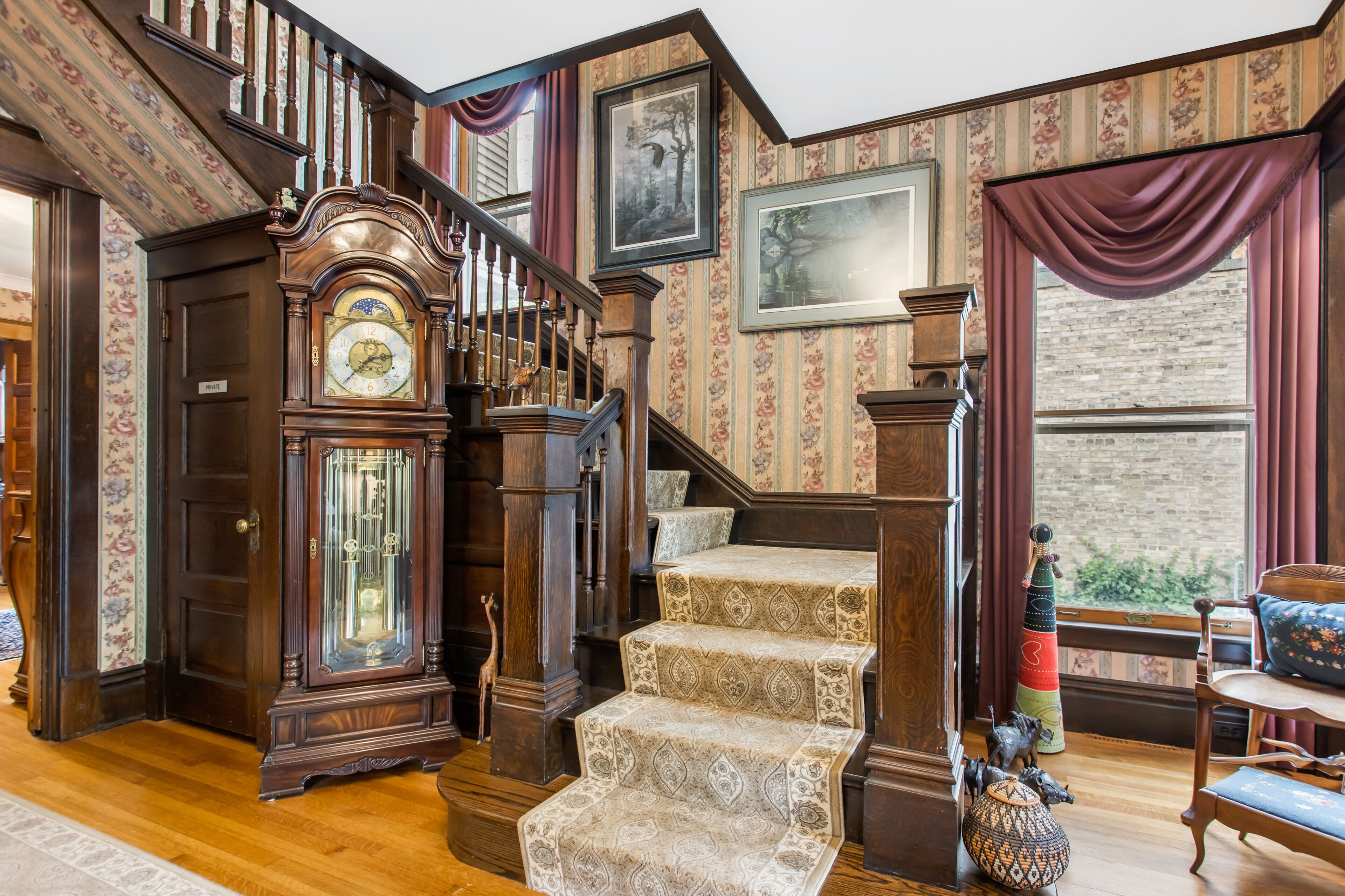 A grand staircase with ornate woodwork, a large grandfather clock, and a cozy seating area with a view of the outdoors.