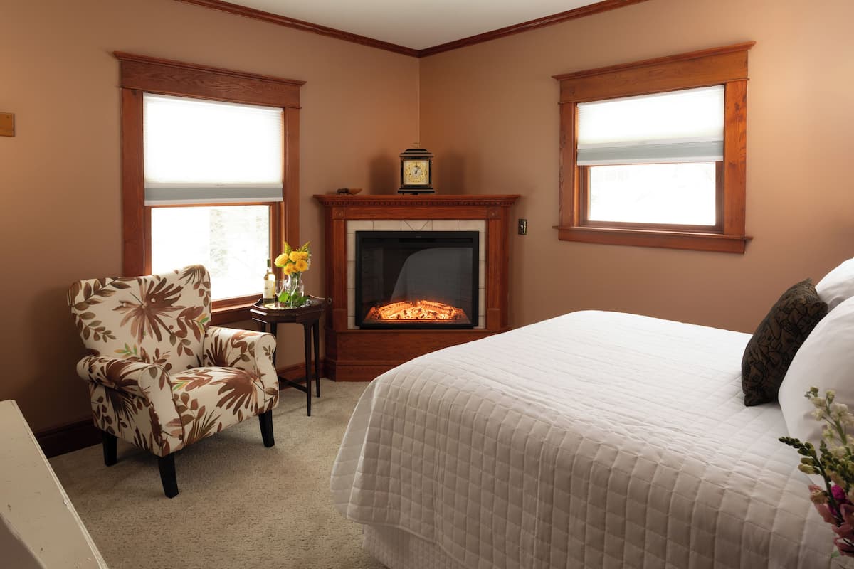 A bedroom with a fireplace, a bed, a chair, and two windows.