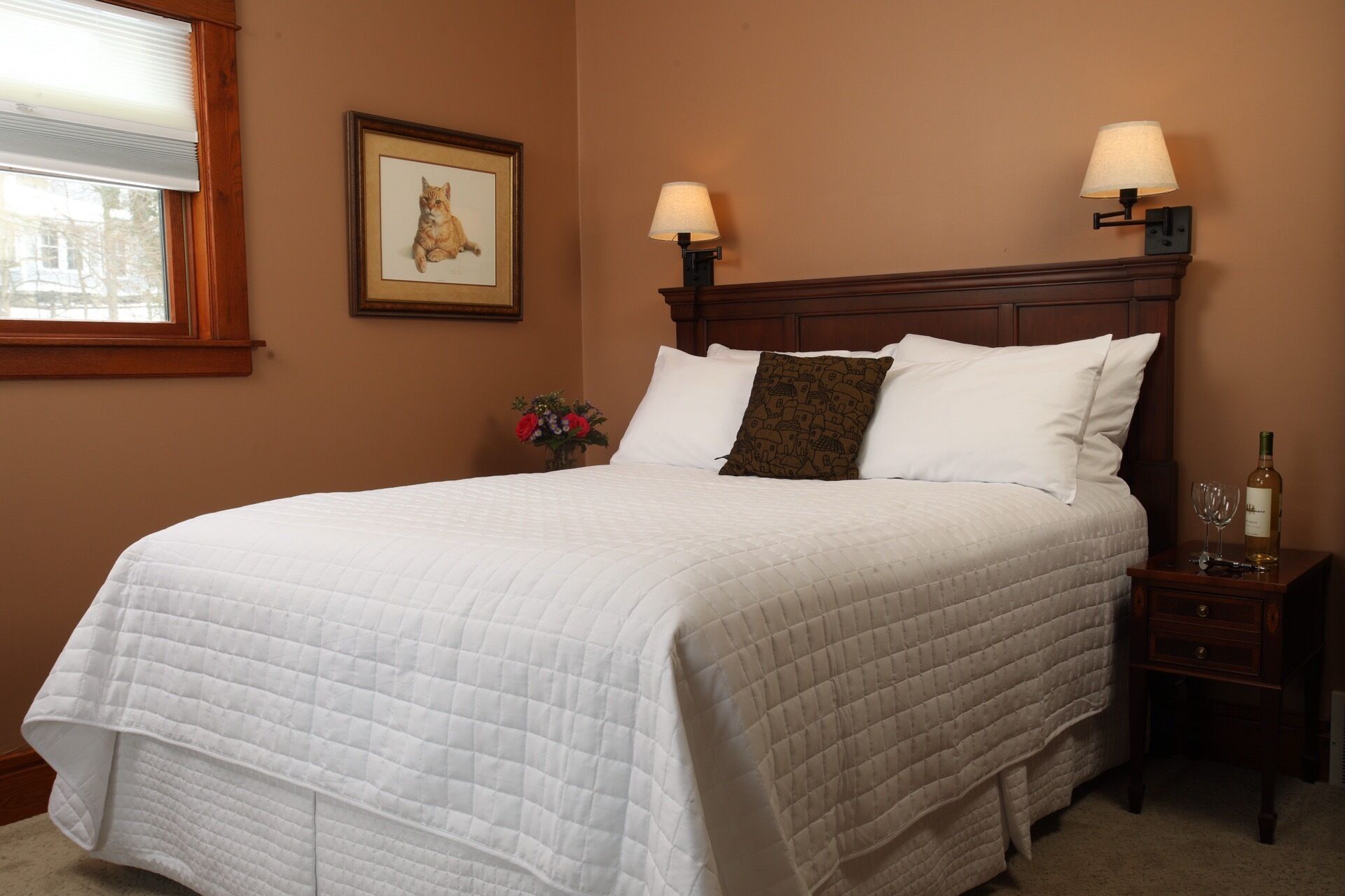 A neatly made bed with a white quilted bedspread, two white pillows, and a brown patterned pillow. There are two wall-mounted lamps on either side of the bed, a framed picture on the wall, and a small table with a bottle and glass to the right.