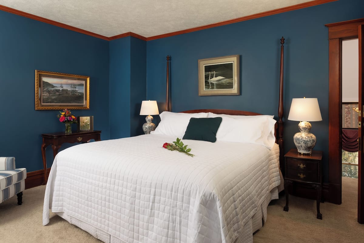 A bedroom with a large bed, two bedside tables with lamps, a nightstand, a framed picture on the wall, and a vase with flowers on the nightstand.