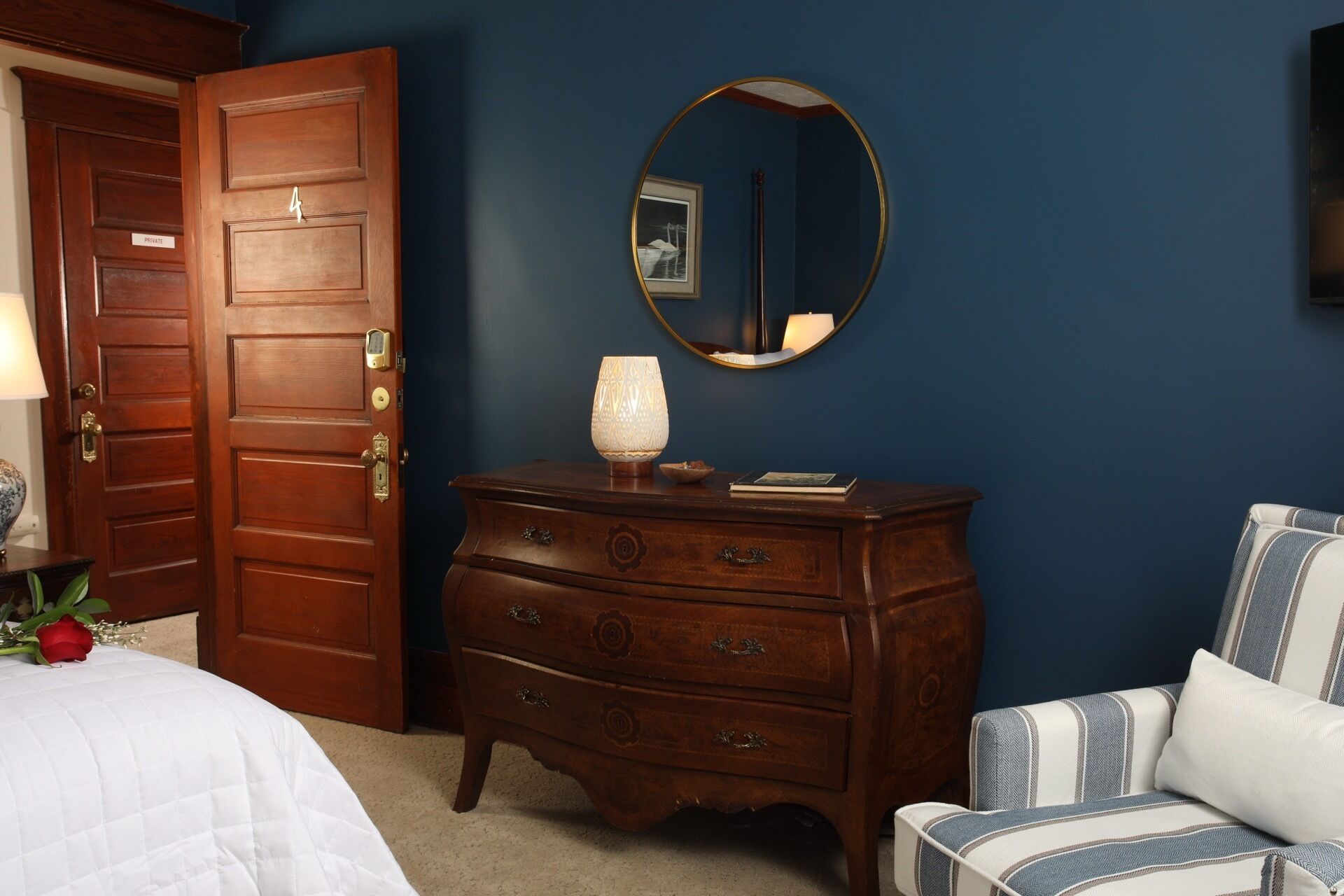 Une chambre avec une commode en bois, une table de chevet avec une lampe, un lit recouvert d’une couette blanche, une chaise à rayures bleues et blanches, et un miroir rond accroché au mur.