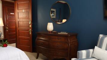 A bedroom with a wooden dresser, a bedside table with a lamp, a bed with a white duvet, a blue and white striped chair, and a round mirror on the wall.