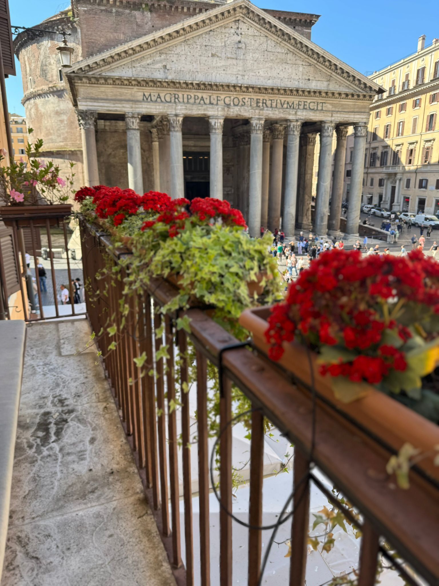 Ein Balkon mit roten Blumen und Blick auf ein klassizistisches Gebäude mit Säulen und Inschriften.