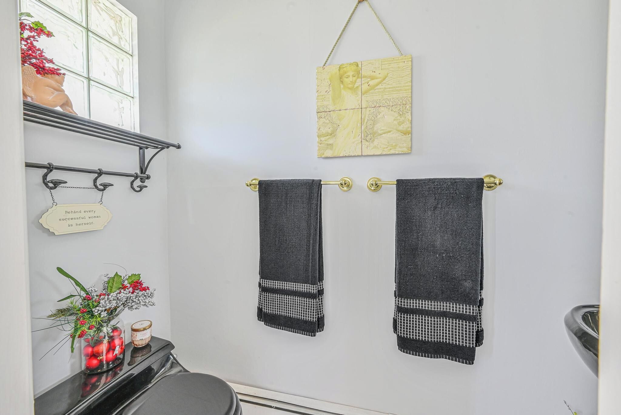 Un baño con dos toallas colgadas en un perchero, un letrero decorativo y un jarrón con flores sobre una estantería.