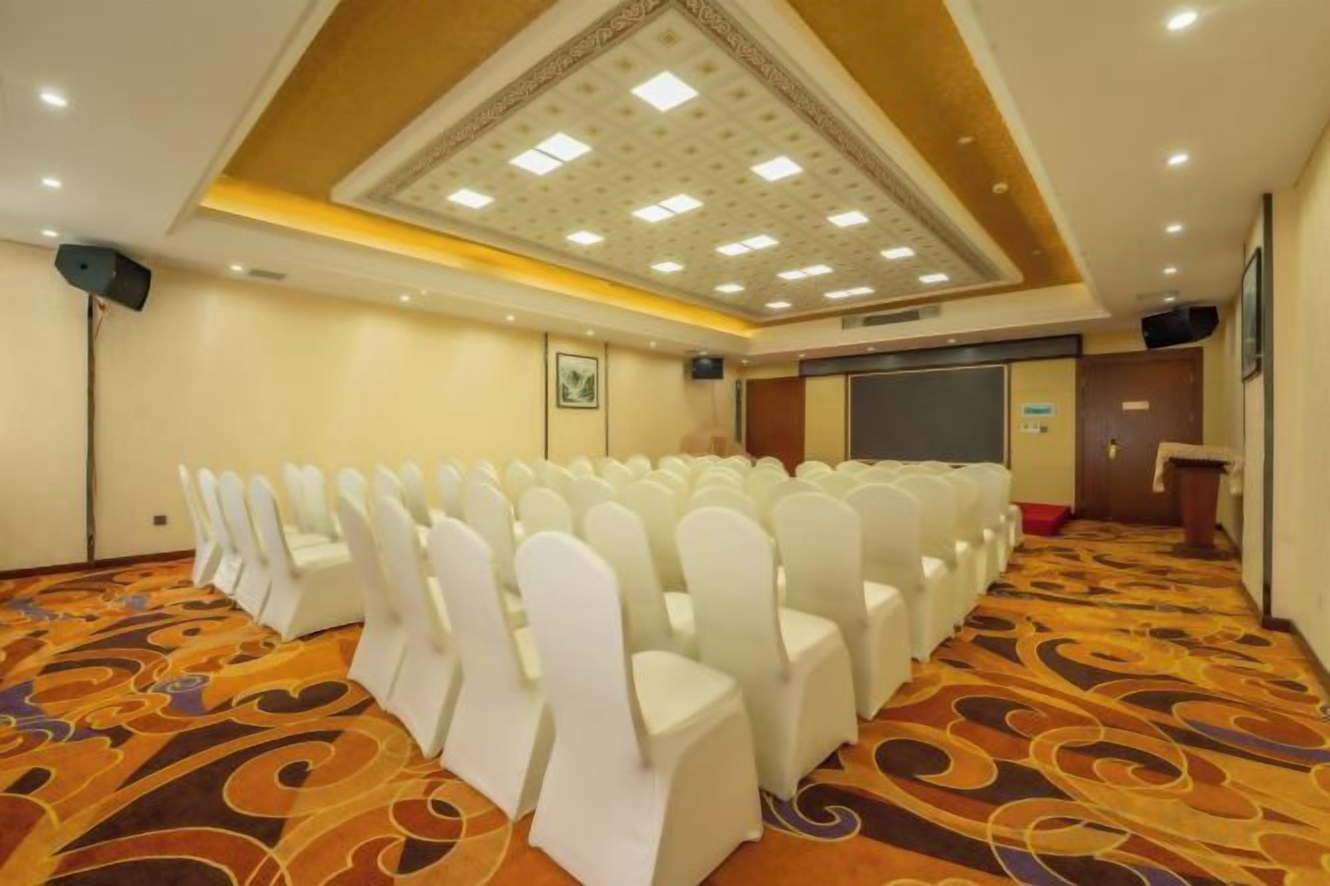 A conference room with rows of white chairs, a carpeted floor, a speaker, and a projection screen.