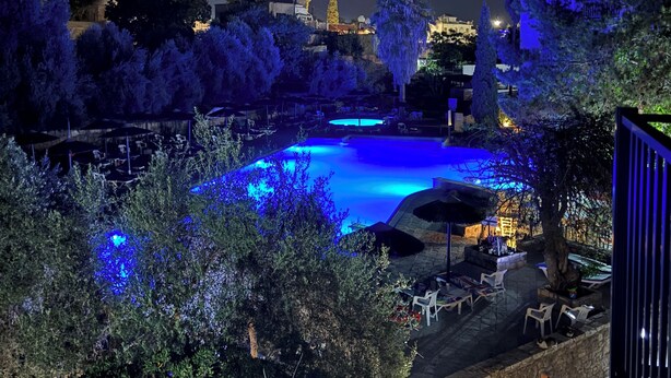 Vista nocturna de una zona de piscina con iluminación azul, rodeada de árboles y edificios.