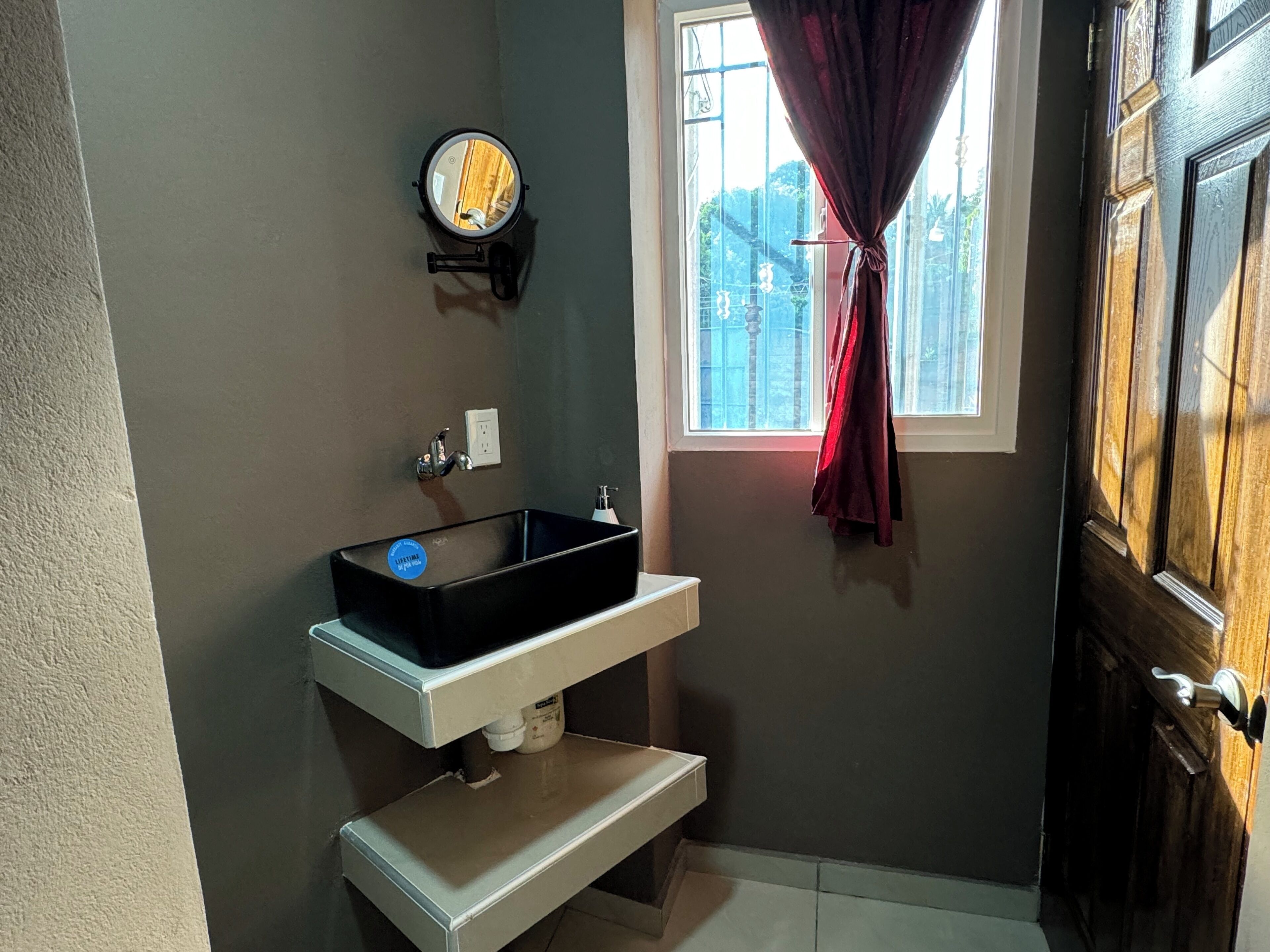Een badkamer met een zwart wastafel, een spiegel, een raam met gordijn, en een deur.