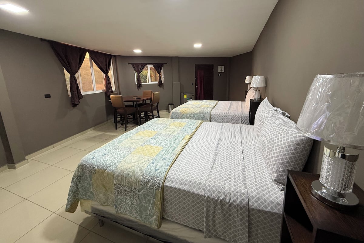 A hotel room with two beds, a dining table, chairs, a lamp, and a window with curtains.