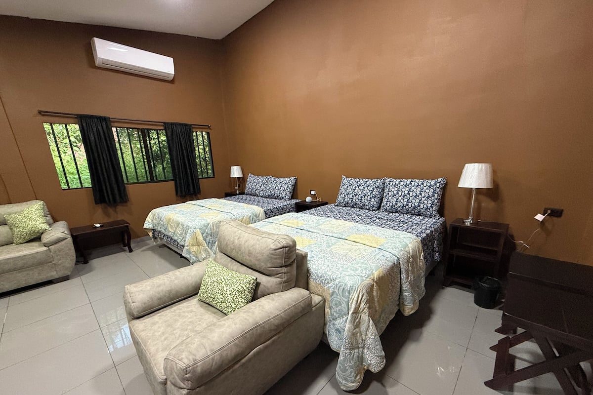 A hotel room with two beds, a sofa, a small table, and an air conditioning unit.