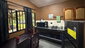 A compact kitchen with a refrigerator, microwave, and coffee maker. A window with curtains is visible, and a small potted plant is on the counter. - El Mangal B&B (Suchitoto)