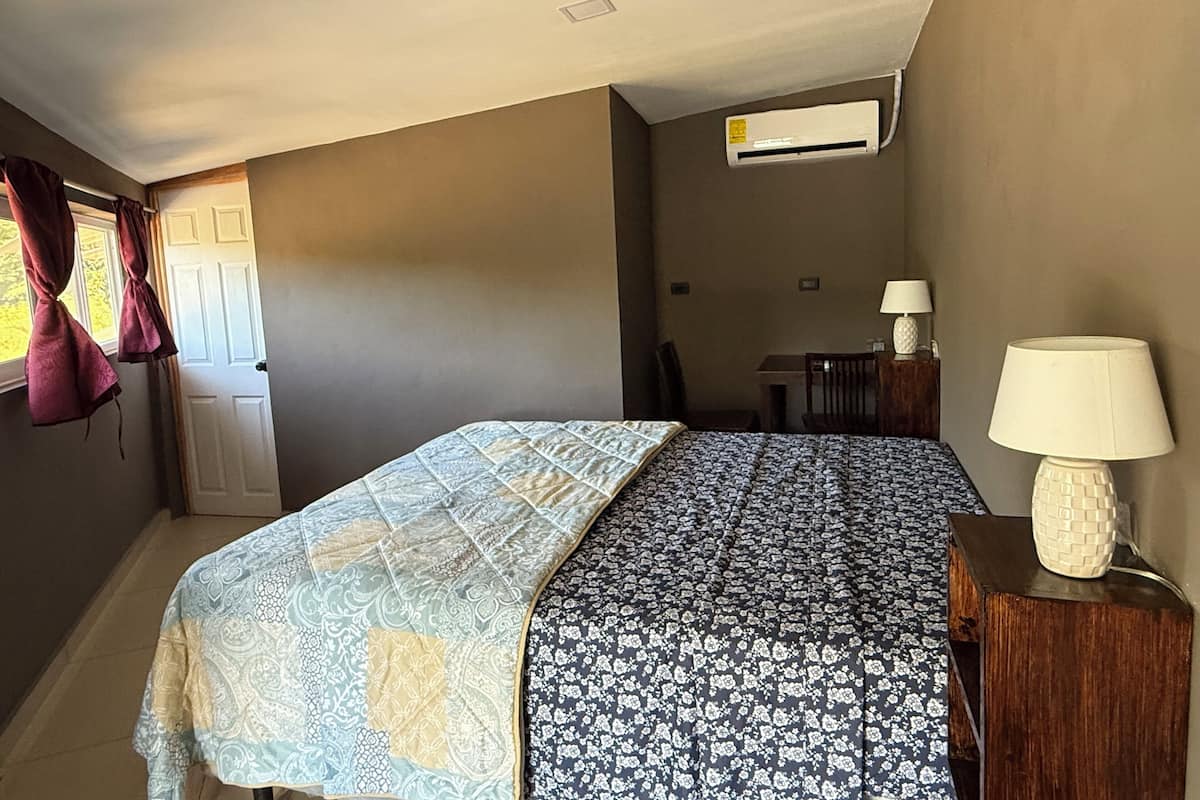A bedroom with a bed, a nightstand with a lamp, a window with curtains, an air conditioning unit, and a small table with a chair.