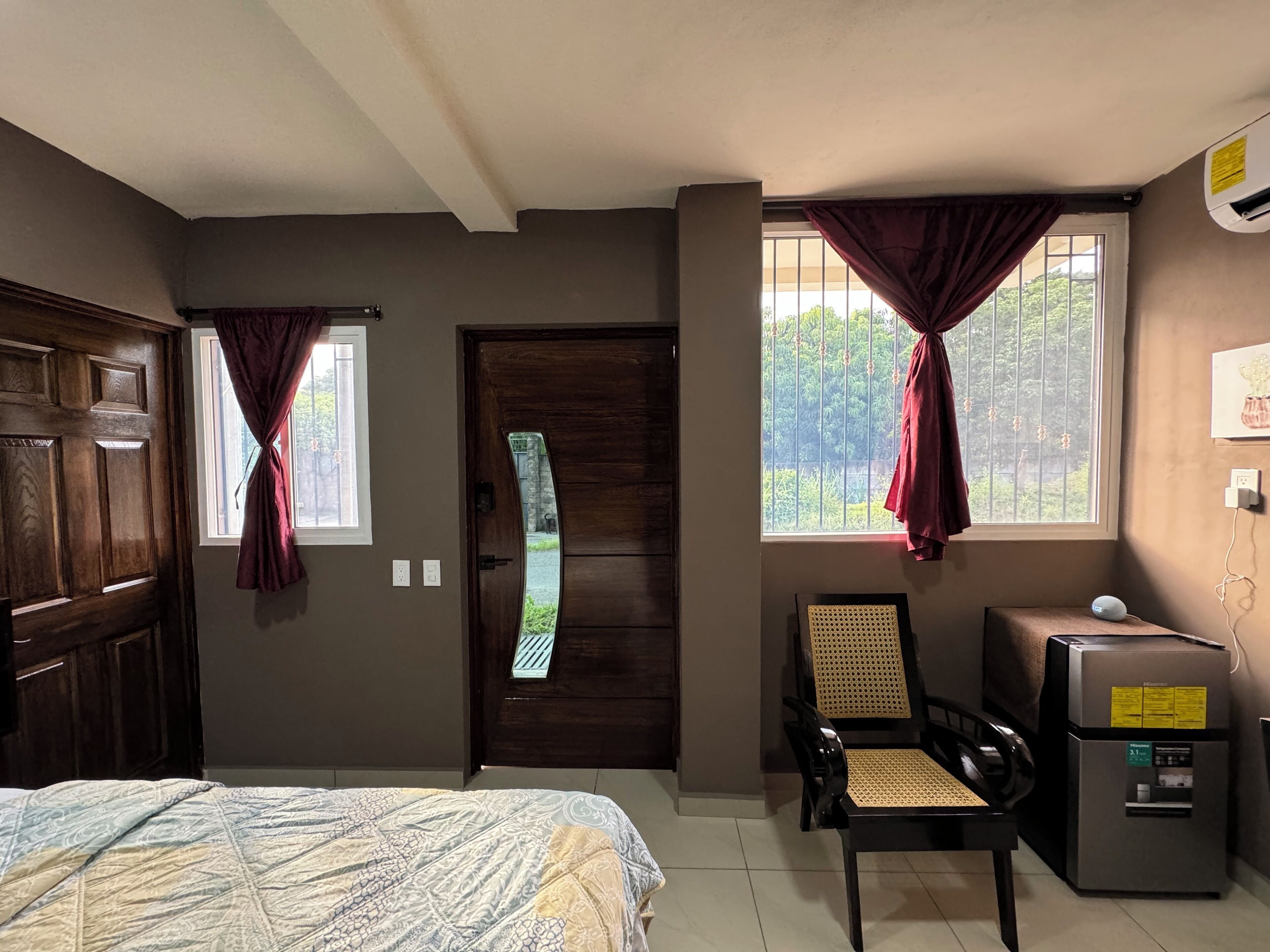 A room with a bed, a chair, a small table, a refrigerator, and two windows with curtains.