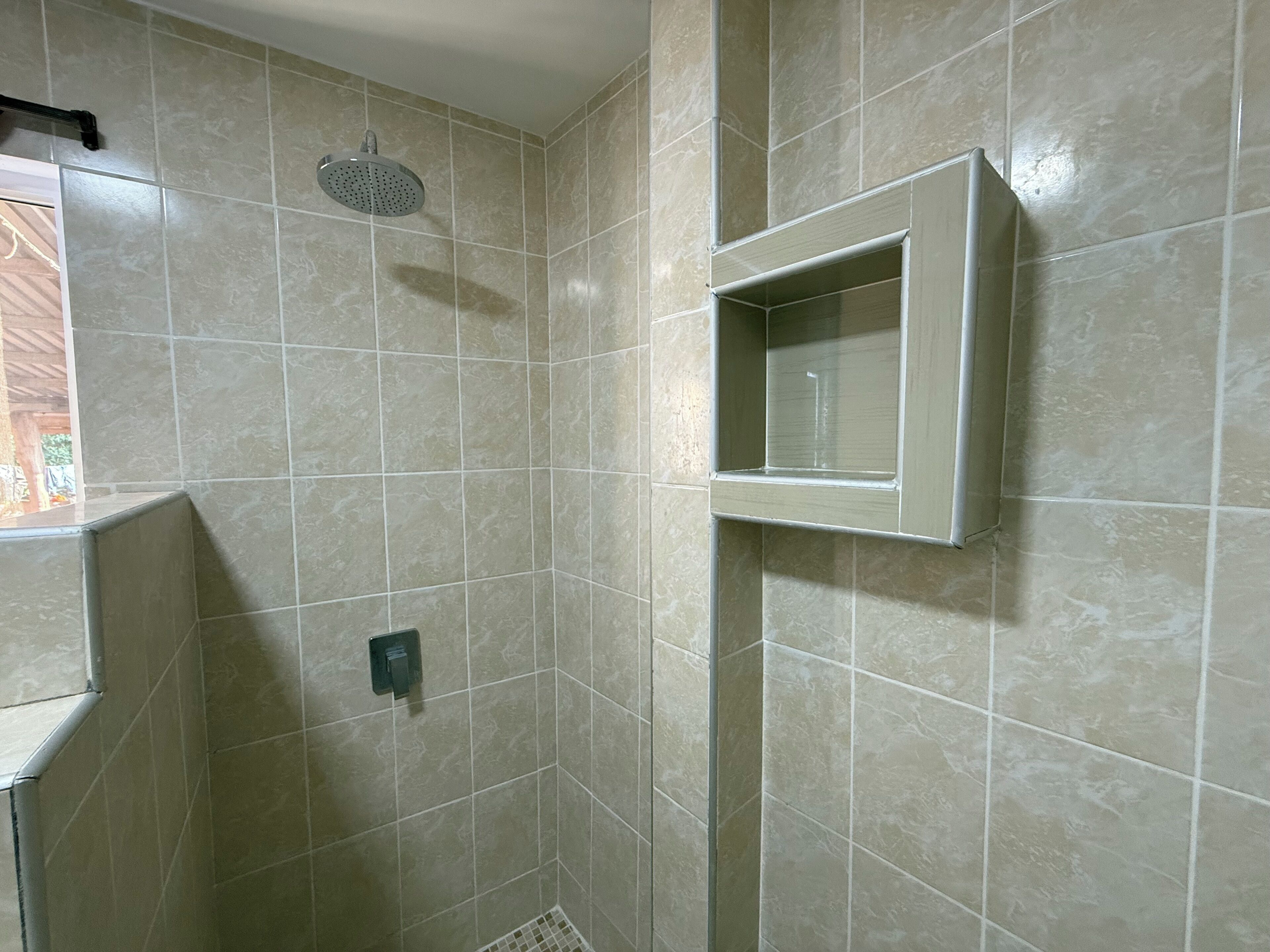Une salle de bain avec des carreaux de couleur beige, une pomme de douche, une petite étagère et une bouche d’aération.