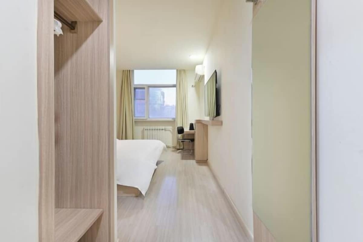 A hotel room with a bed, a desk, a chair, a radiator, and a window.