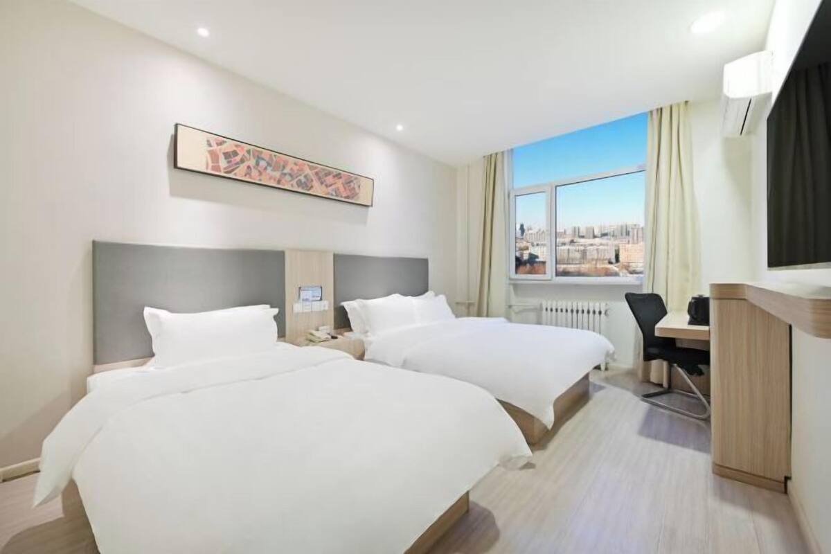 Habitación de hotel con dos camas, un escritorio, una silla, ventana con vista a la ciudad y un cuadro en la pared.